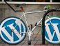 Bicycle with WordPress logos on wheels locked to bike rack outside building.