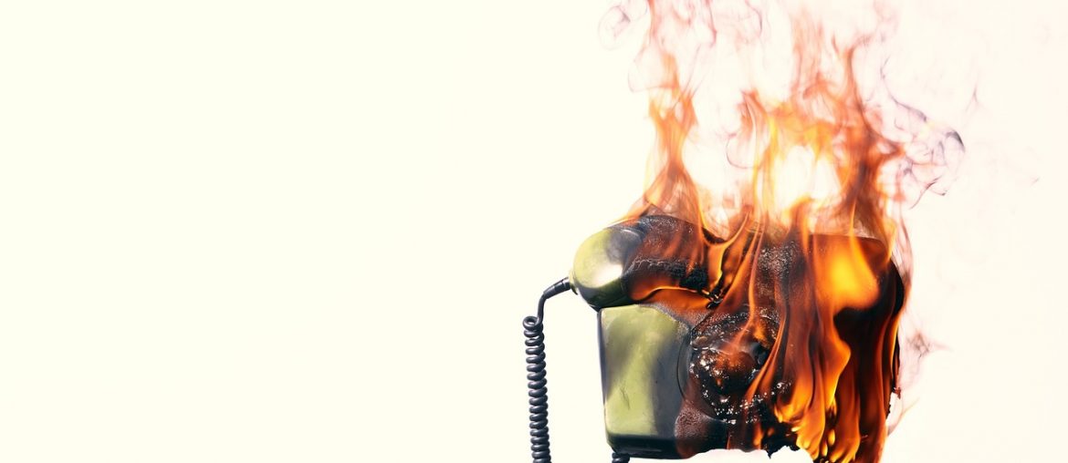 Vintage rotary telephone engulfed in orange flames with rising smoke against a white background.
