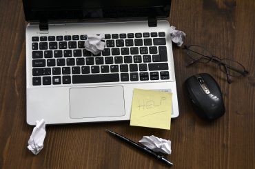 Laptop with crumpled paper, a sticky note with Help, glasses, a pen, and a mouse on a wooden desk.