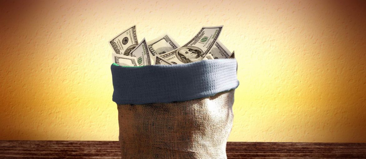 A sack overflowing with US dollar bills on a wooden table against a warm, textured yellow background.