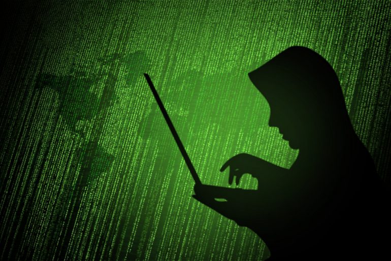 Silhouette of a person in a hoodie typing on a laptop against a backdrop of green binary code and digital world map