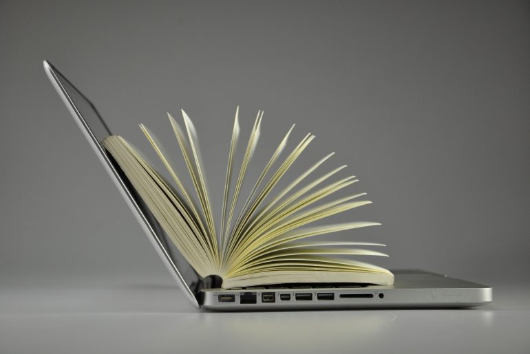 Open laptop with a book standing upright on the keyboard, pages fanned out, representing technology and education.