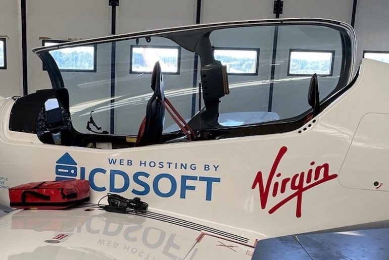 Cockpit of a small white aircraft with Virgin and ICDSoft branding inside a hangar, featuring large side windows.