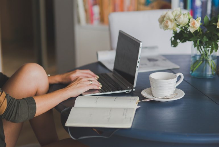 Person typing on a laptop at a table with a notebook, coffee cup, and vase of white roses in a cozy home setting.