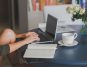 Person typing on a laptop at a table with a notebook, coffee cup, and vase of white roses in a cozy home setting.